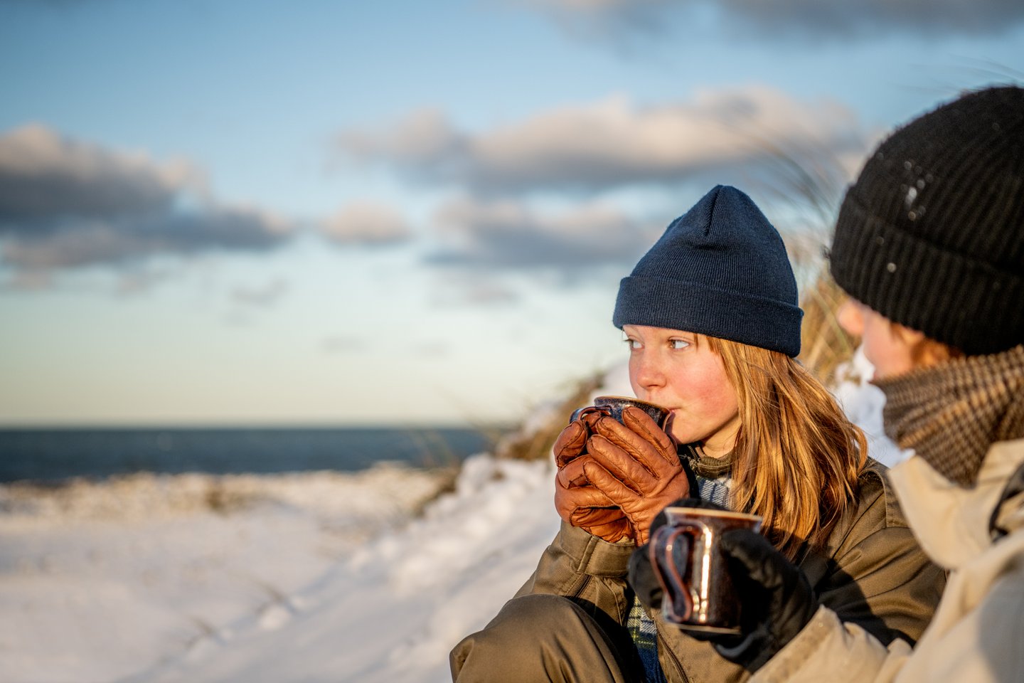 Jente holder varmen med en kopp varm drikke ved Klitmøller strand om vinteren